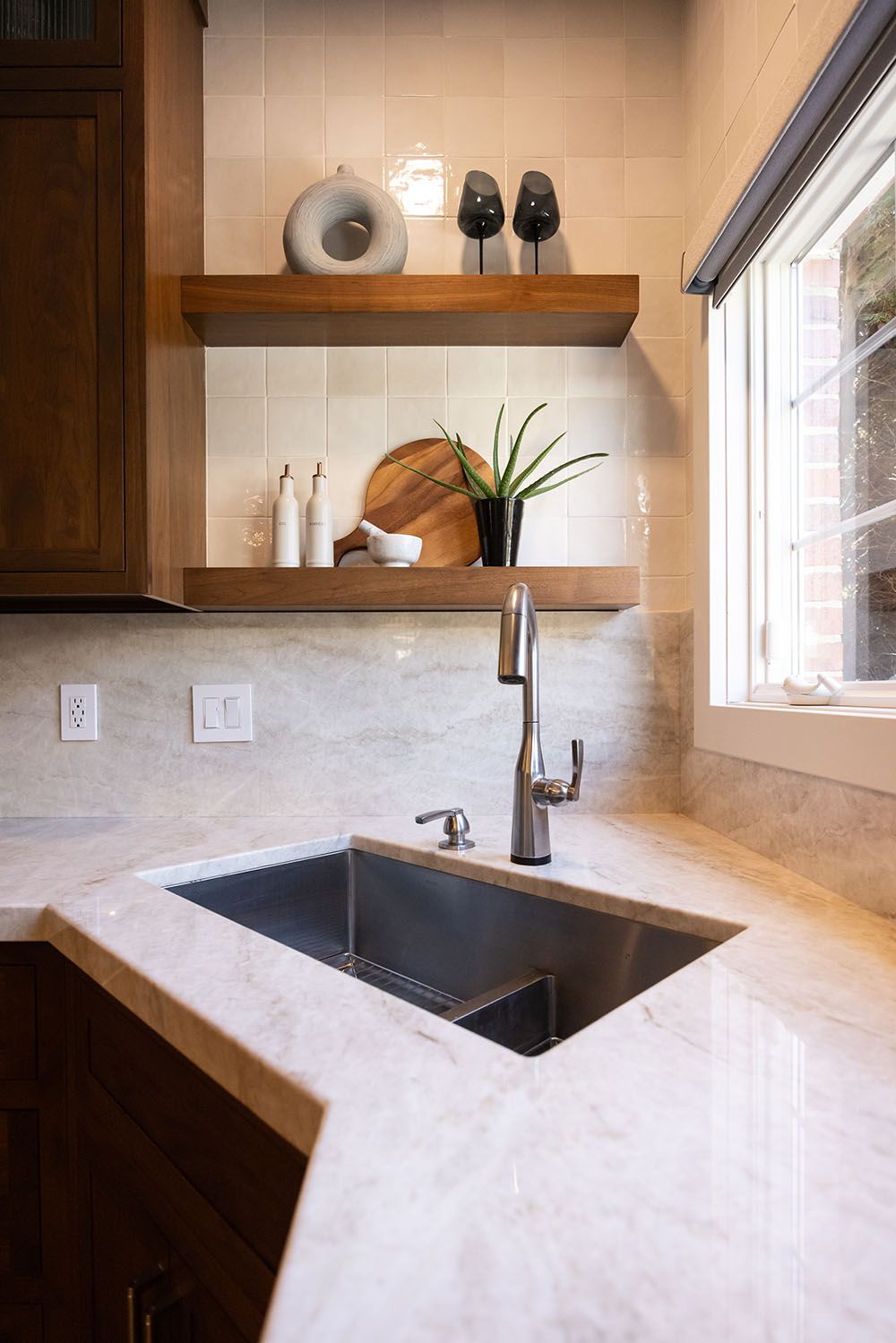 A kitchen sink with a chrome faucet set in a light marble countertop below two floating wooden shelves on tiled walls.