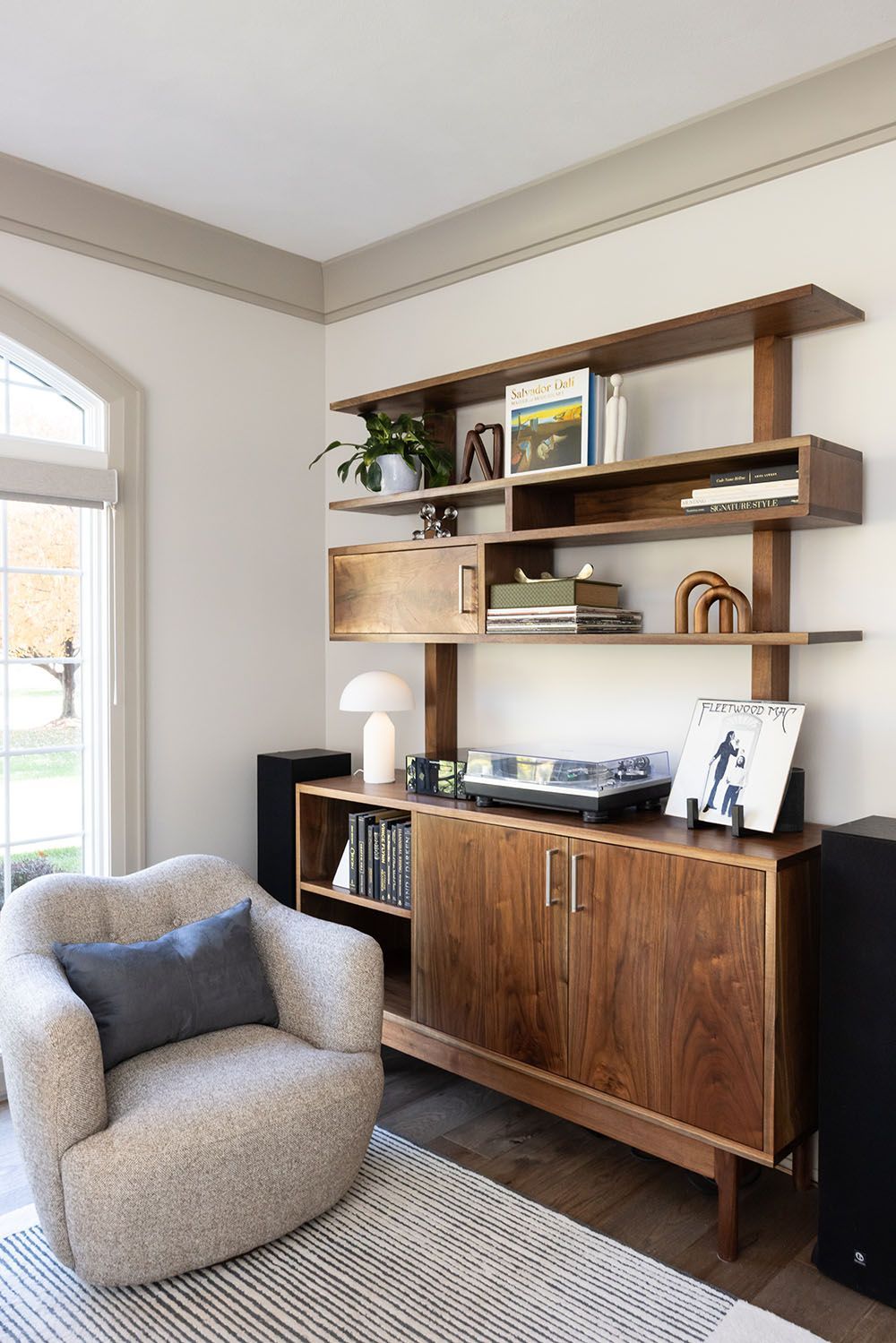 A mid-century modern wooden shelving unit and media console stand next to a textured beige armchair in a bright room.