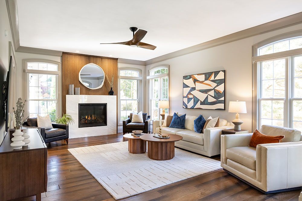 A modern living room with a fireplace, white sofa, armchairs, two round wooden coffee tables, and a large wall mirror.