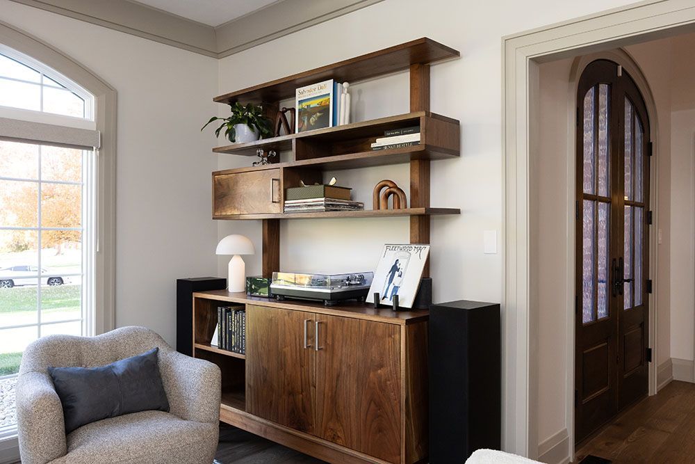 A mid-century modern wooden shelving unit holding a record player and books next to a cream chair and arched doorway.