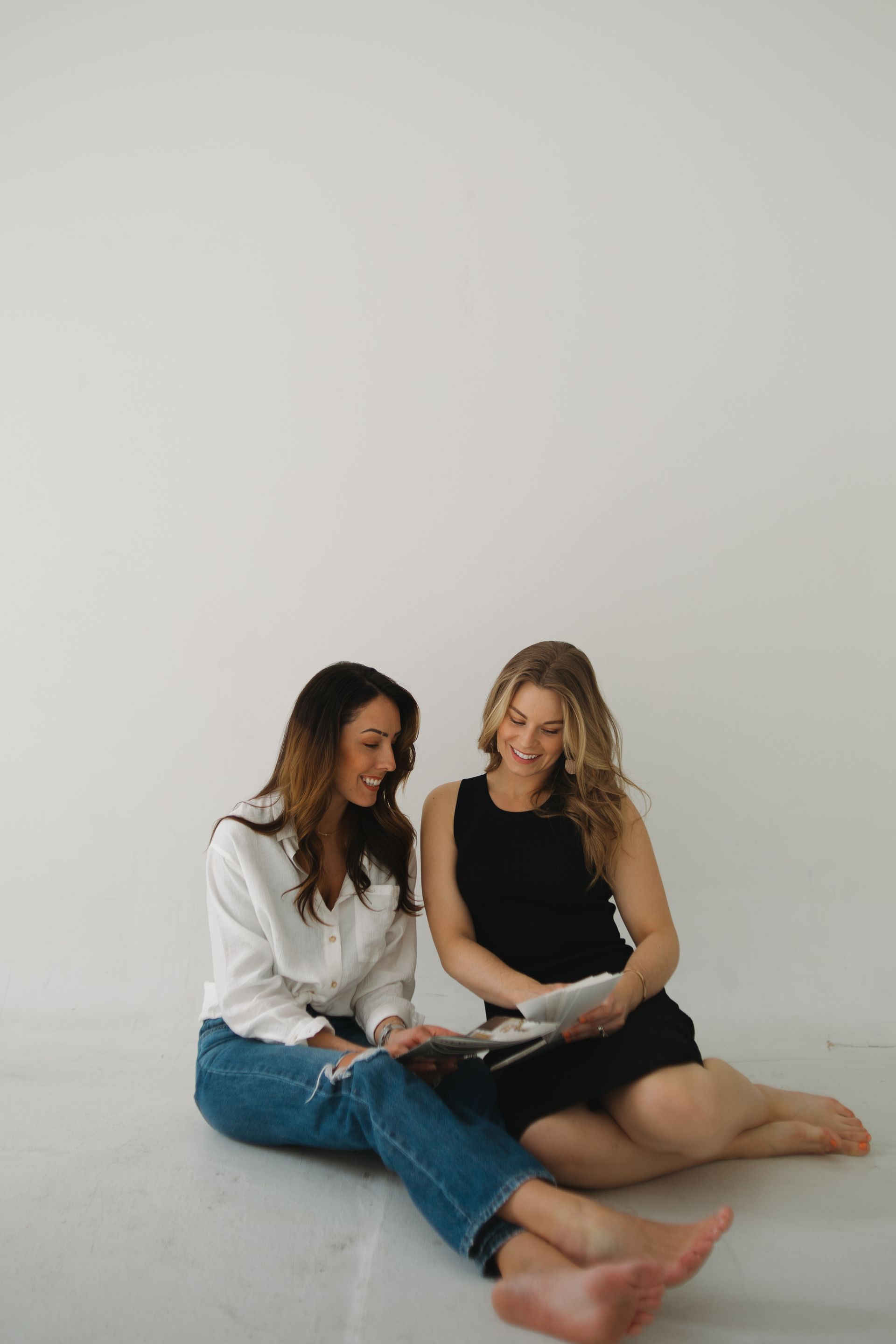 Two women sit on the floor, smiling, looking at a tablet. White wall background.