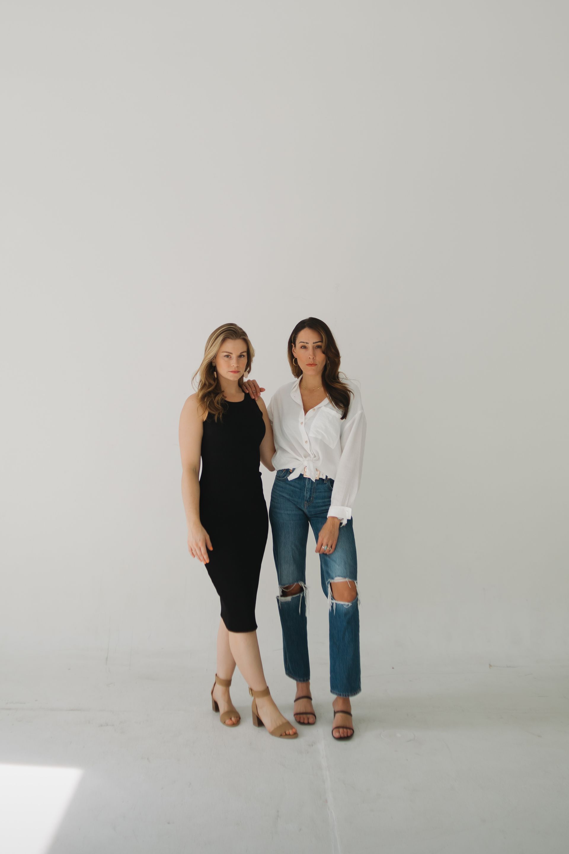 Two women posing in front of a white backdrop. One wears a black dress, the other a white top and jeans.