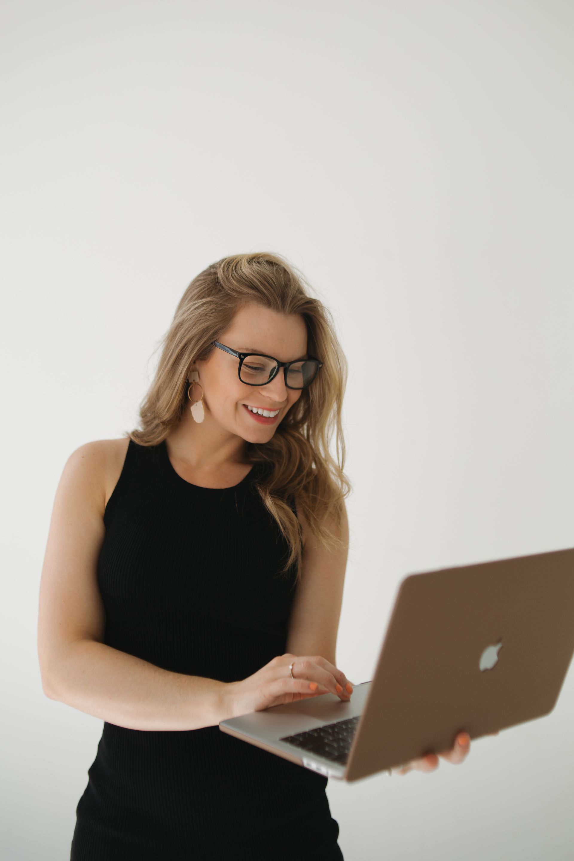 Woman in black dress and glasses smiling while using a laptop in front of a white background.