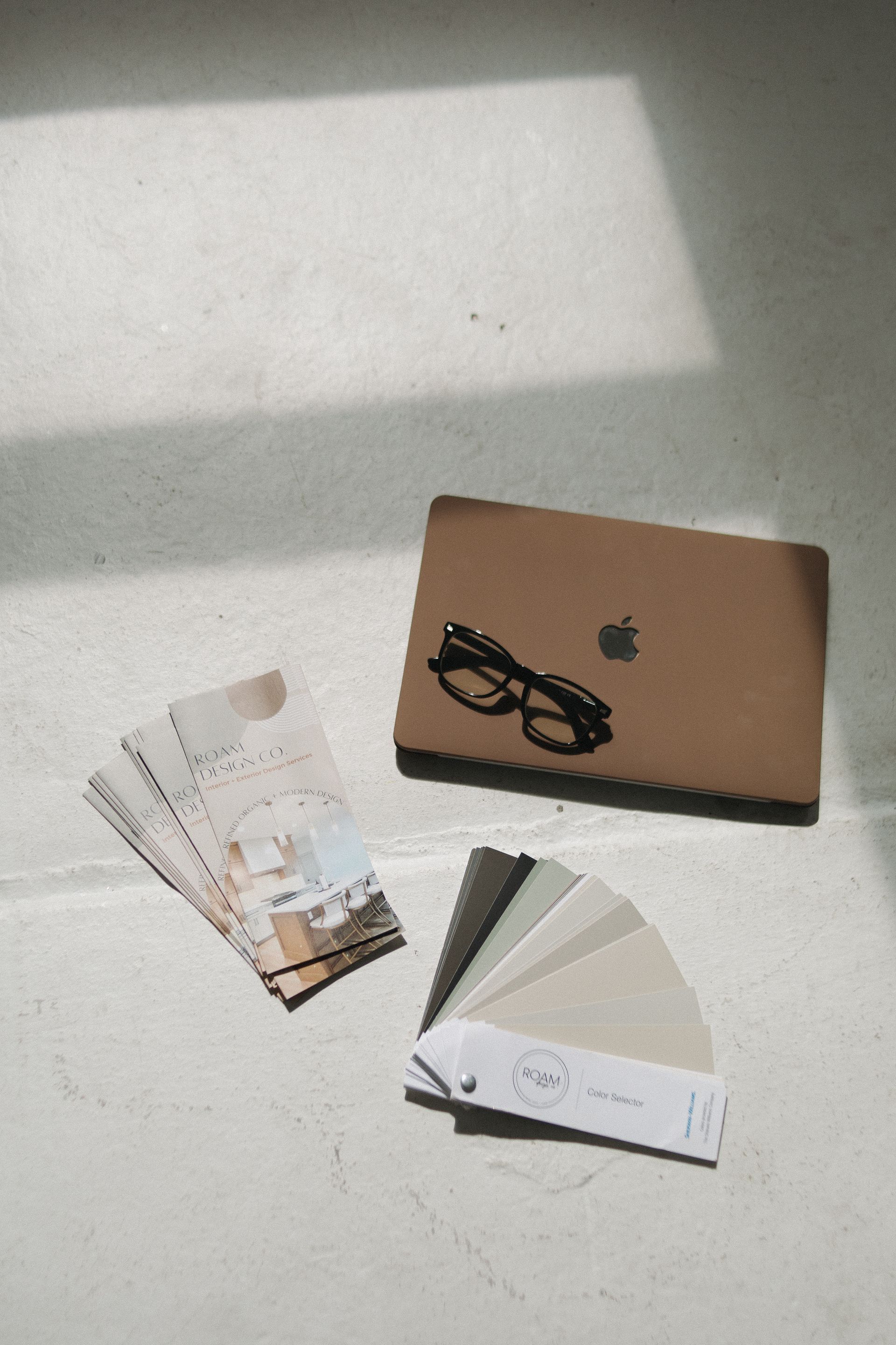 Laptop, glasses, and paint swatch cards on a white surface, bathed in sunlight.