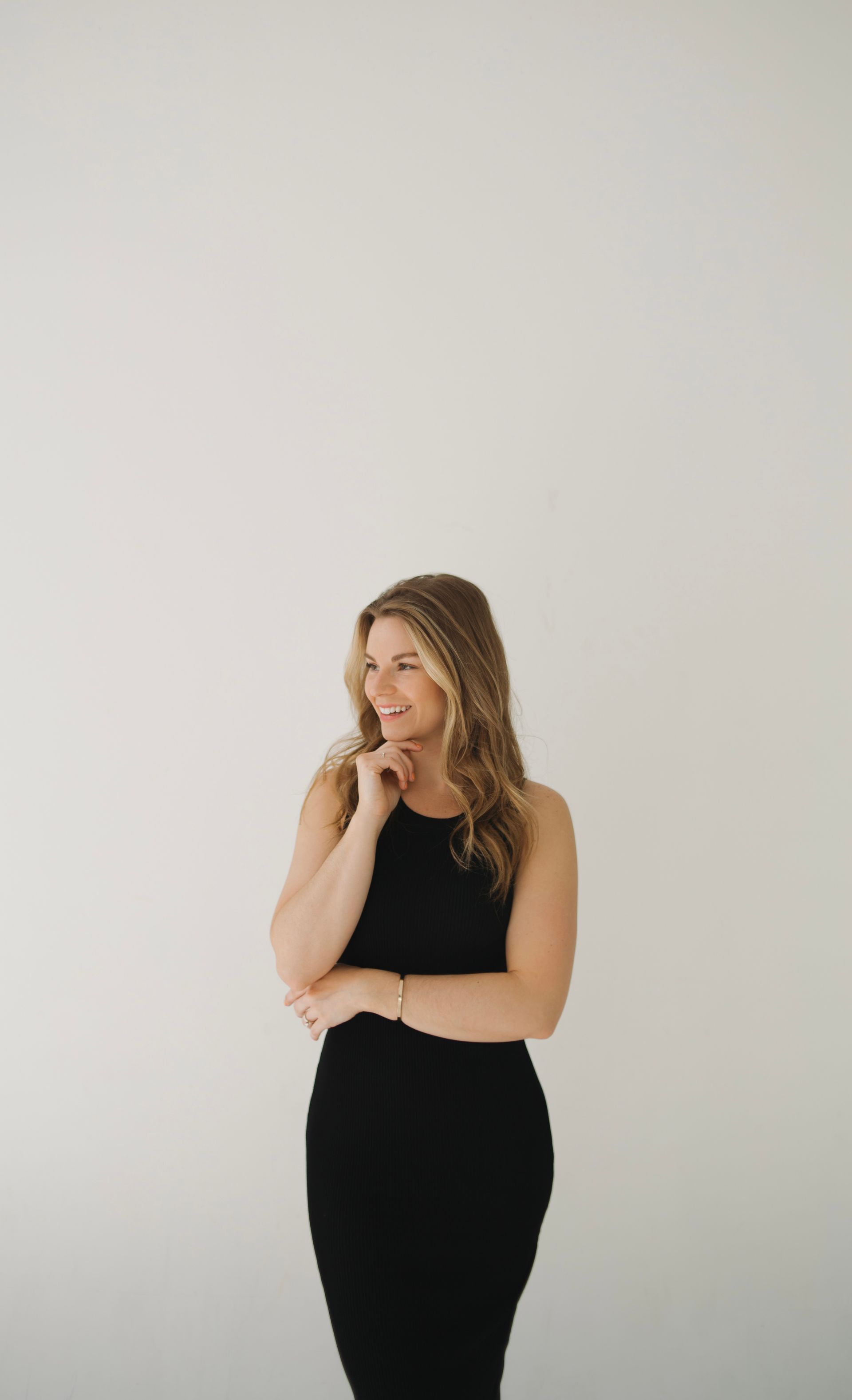 Woman in black dress, smiles thoughtfully with hand on chin, against a white wall.