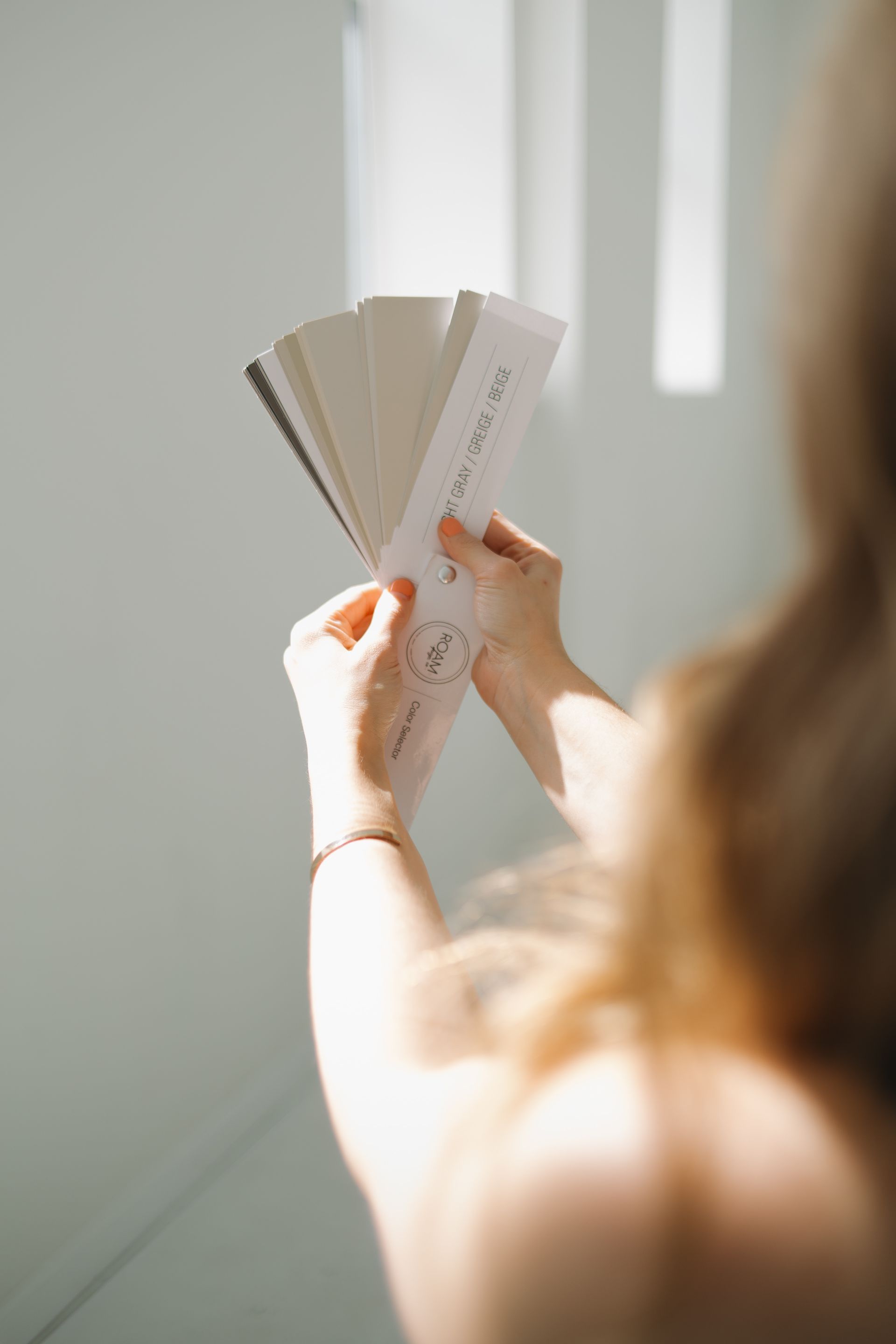 Person holding a paint swatch fan in front of a white wall, comparing colors.