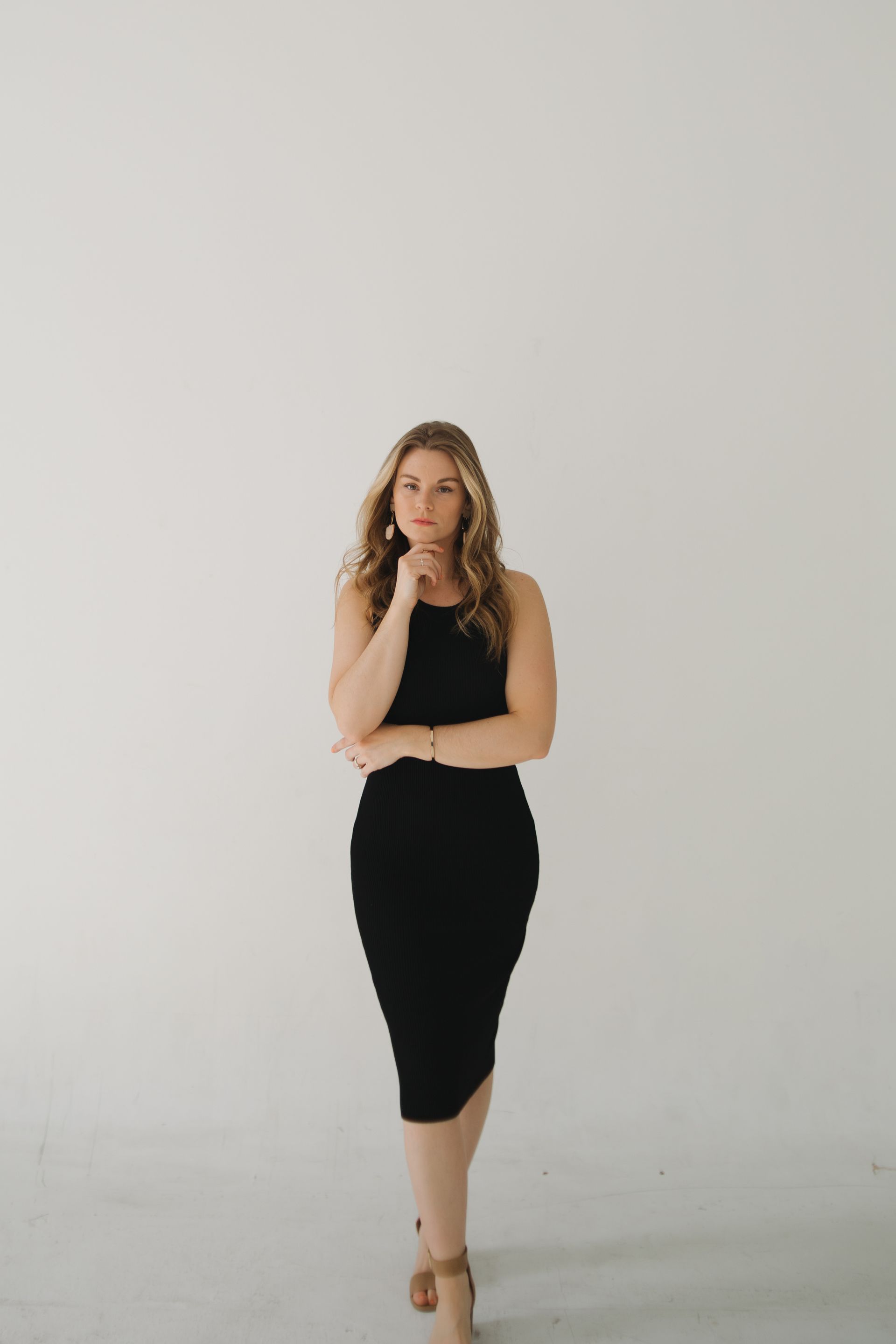 Woman in black dress, standing against a white backdrop, hand to chin, thoughtful expression.