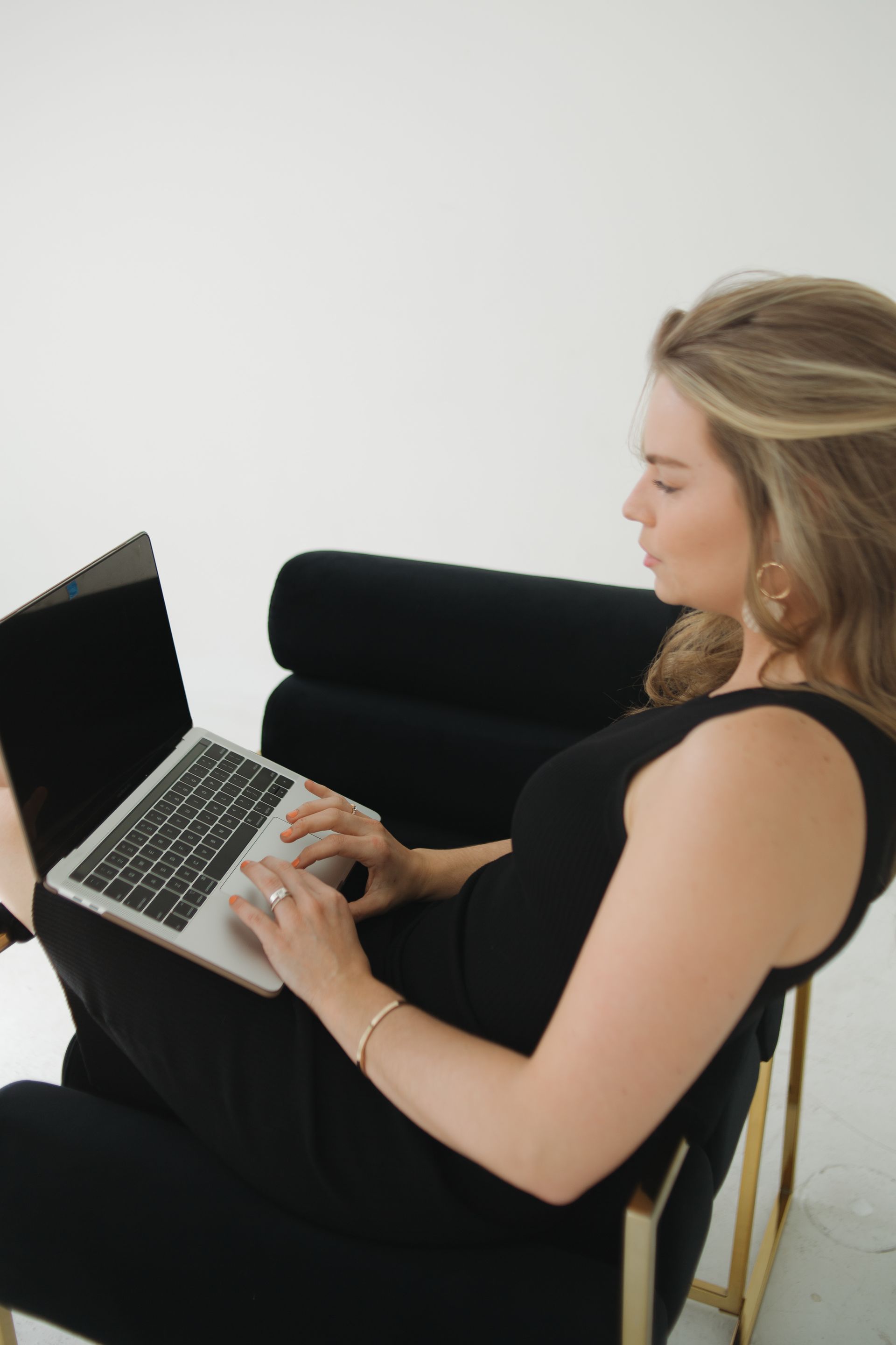 Woman seated in a black chair, using a laptop, in a white-walled room.