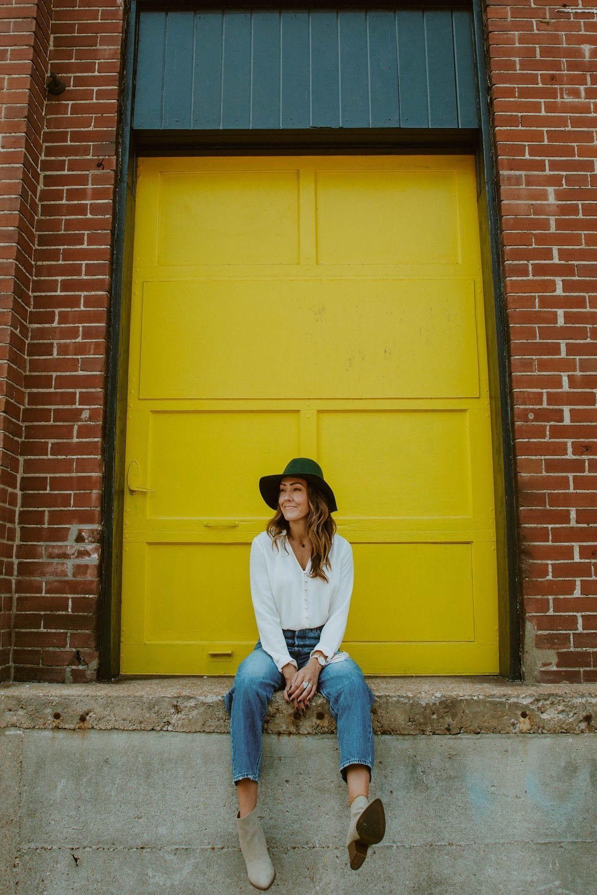 Woman in jeans and a green hat sits in front of a bright yellow door against a brick wall.
