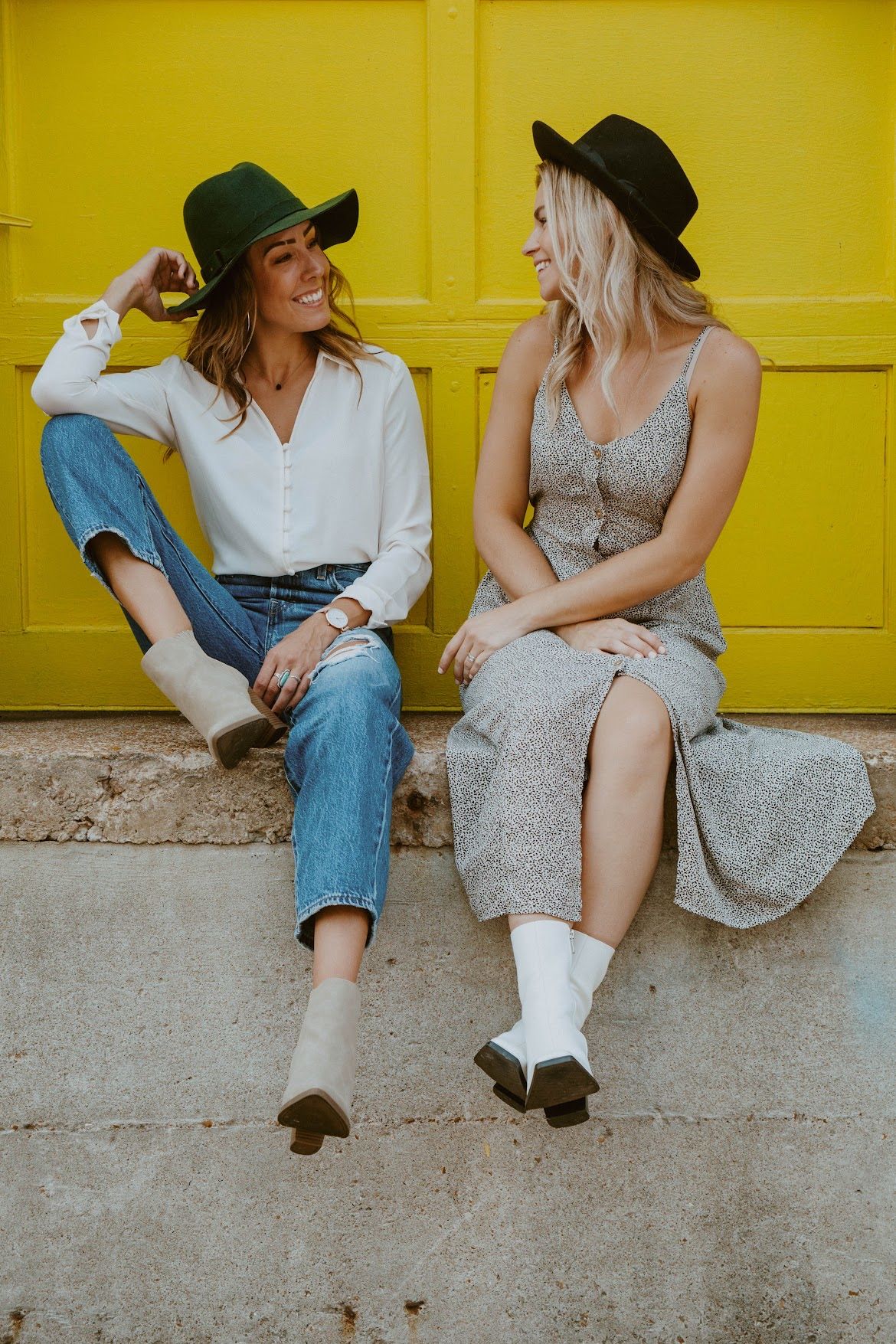 Two women in hats smiling, sitting against a yellow wall, one in jeans, the other in a floral dress.