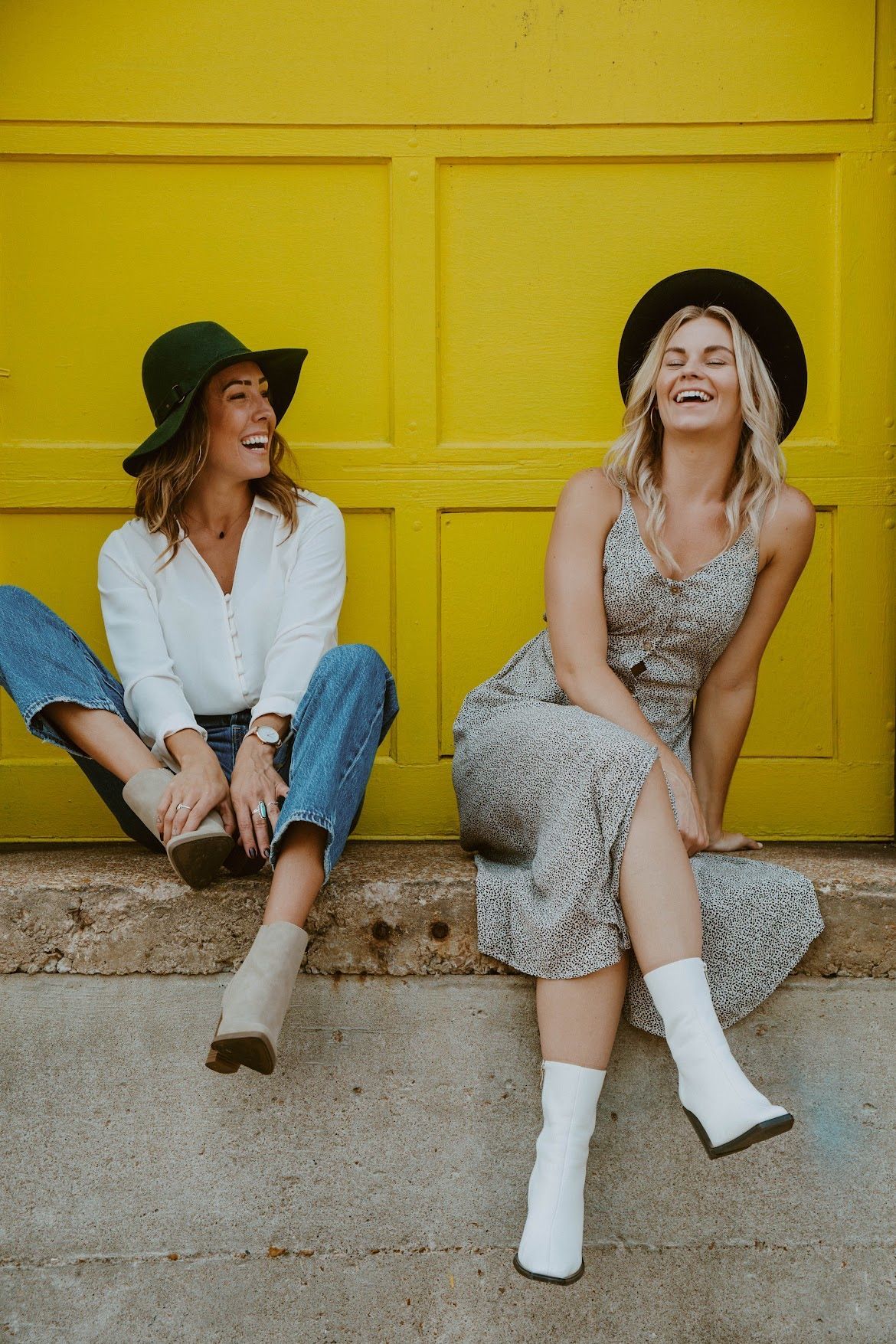 Two women laughing, seated in front of a yellow wall. One wears a hat, jeans, and a white shirt. The other wears a floral dress and a black hat.