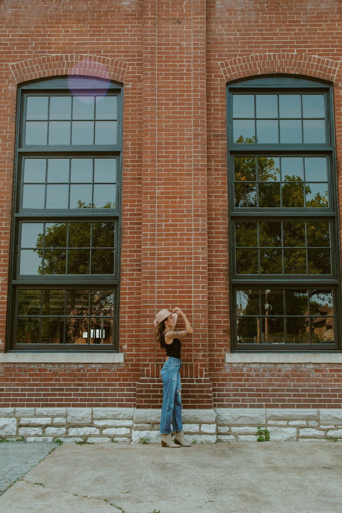 Person in front of a brick building with large windows, wearing a hat, black top, and blue jeans, looking up.