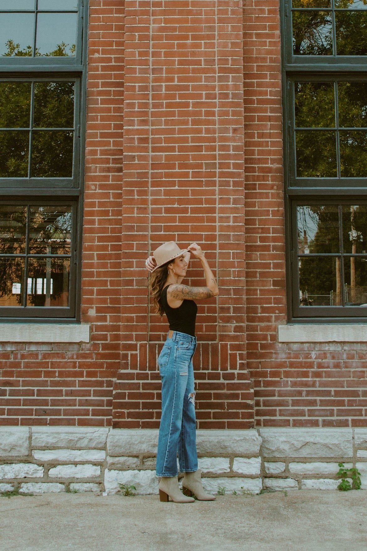 Woman wearing hat and jeans stands against a brick building.