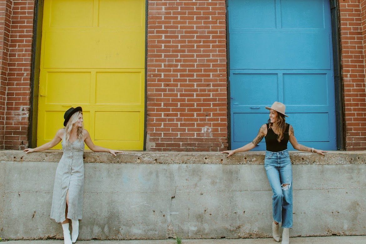 Two women in hats lean on a wall, arms outstretched, beside a yellow and blue door in front of a brick building.
