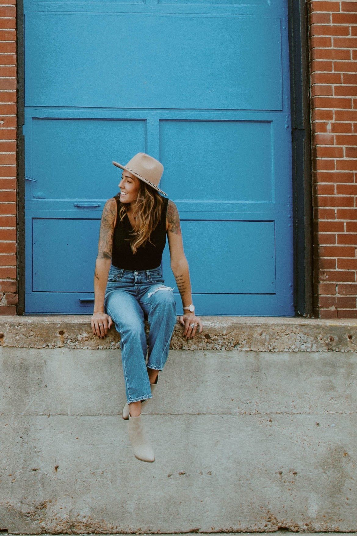 Woman wearing a hat and jeans sits on a concrete ledge, looking to the side, blue door background.