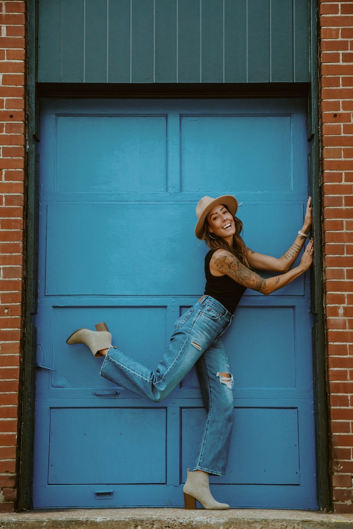 Woman in hat and ripped jeans smiles, leaning against a blue door in a brick building.