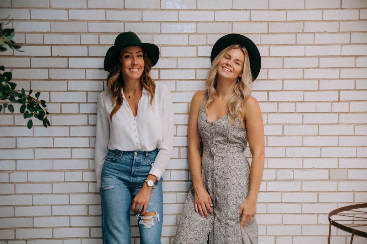 Two women pose against a white brick wall; one in jeans and white shirt, the other in a patterned dress.