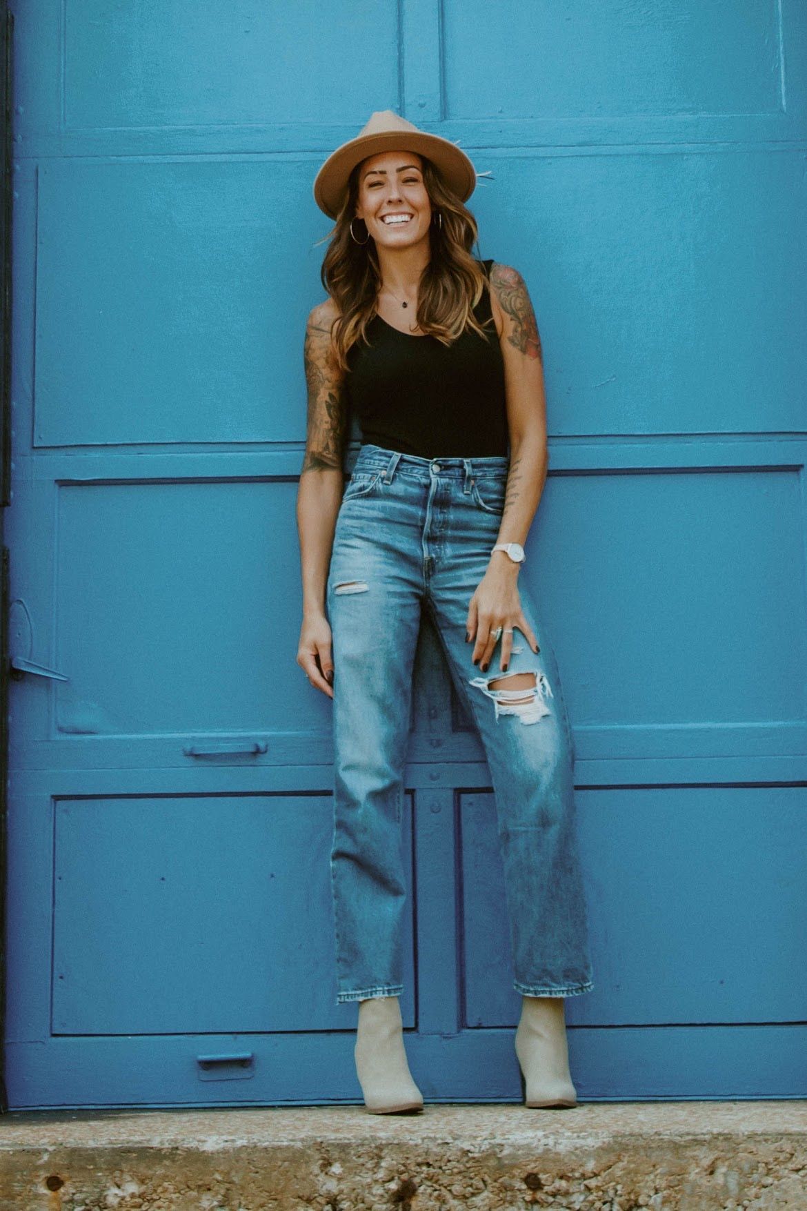 Woman in a hat and ripped jeans smiles in front of a blue door.