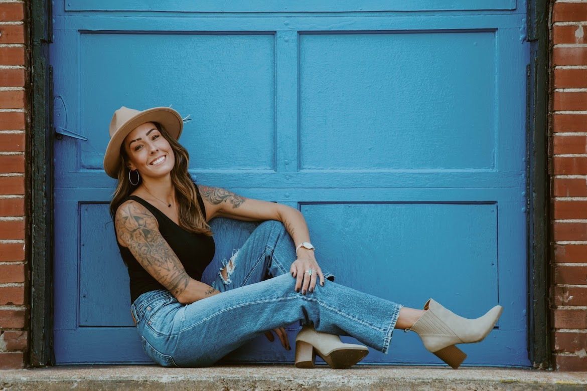 Woman in hat, jeans, and boots, smiling, leaning against a blue garage door, brick wall background.