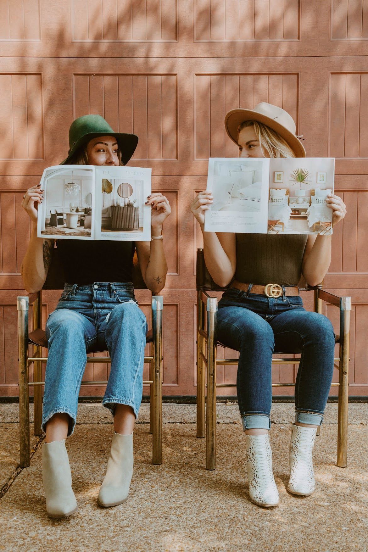 Two people sitting, holding open magazines. They wear hats, jeans, and boots, in front of a closed red garage door.