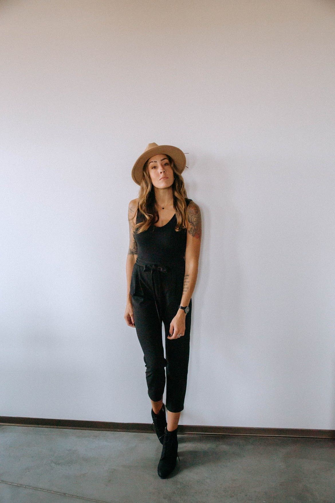 Woman in black jumpsuit, hat, and boots leans against a white wall in a room.