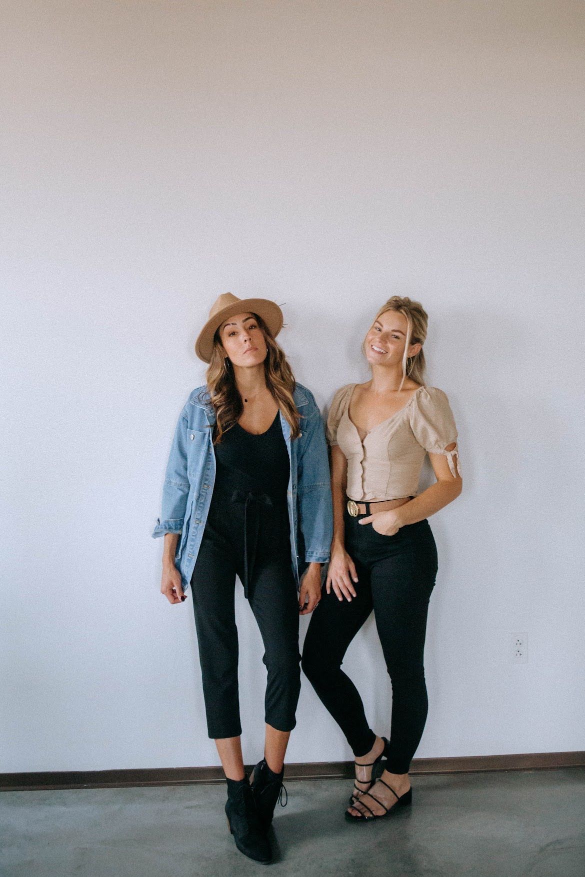 Two women posing against a white wall. One wears a denim jacket and hat, the other a tan top.