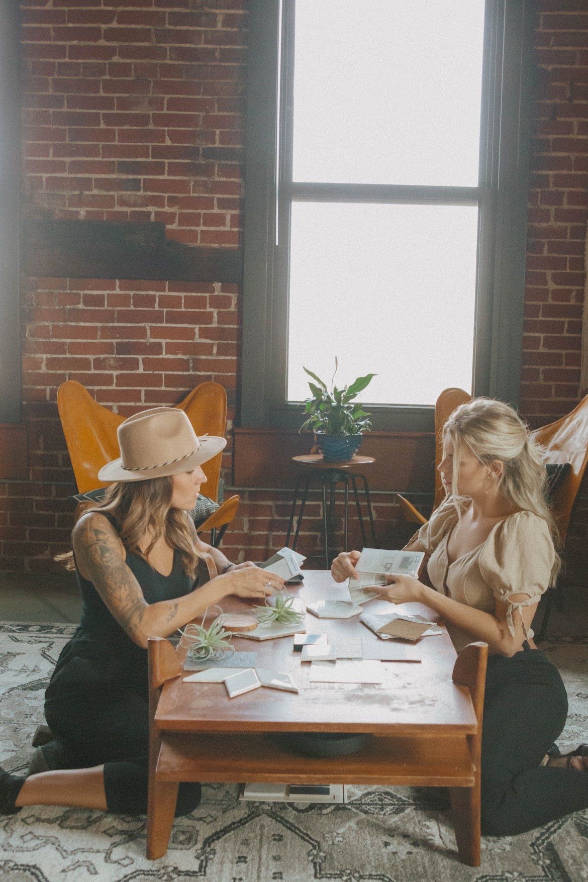 Two women seated at a table, playing cards. Exposed brick wall, large window, bohemian style.