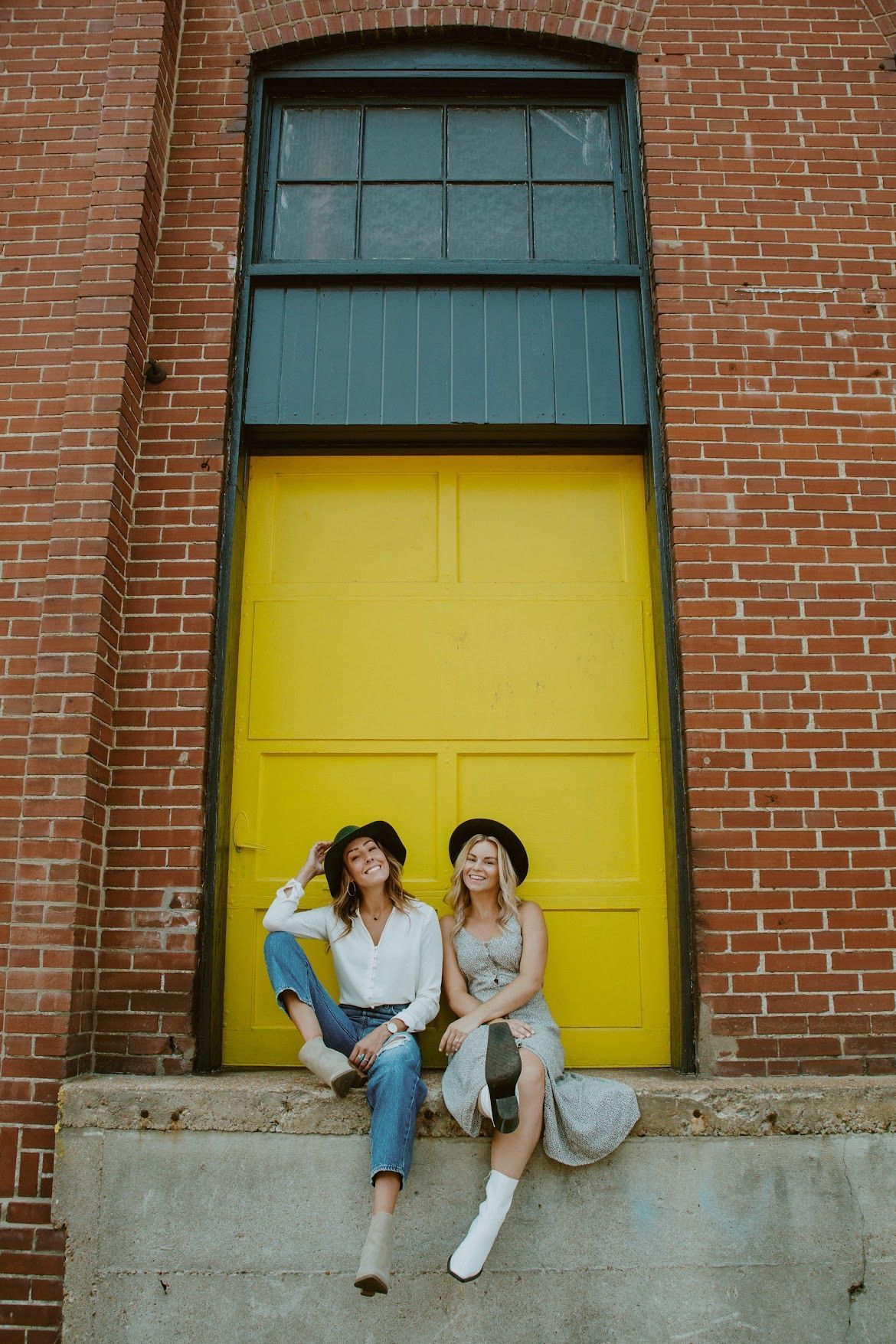 Two people in hats sitting on a concrete ledge in front of a yellow door within a brick building.