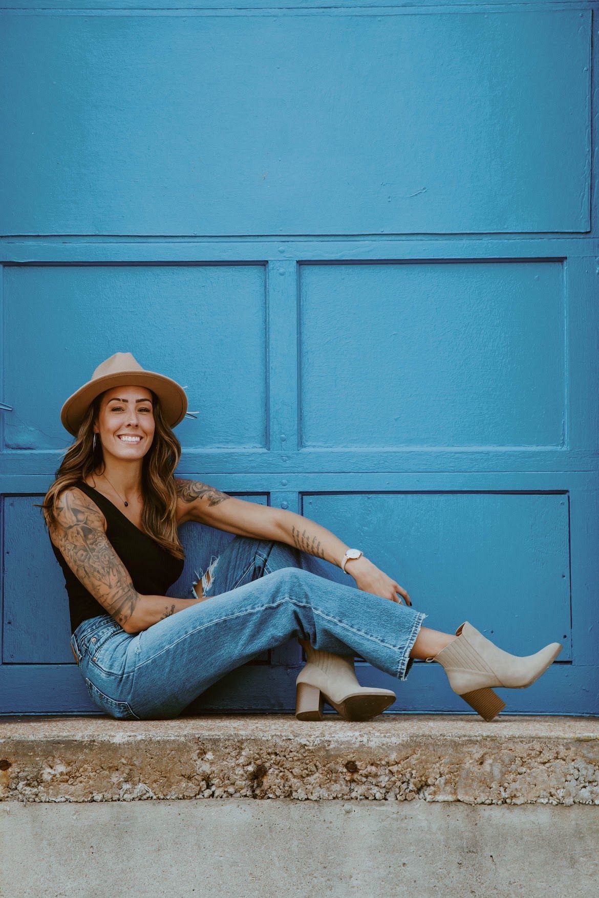 Woman in hat and jeans, smiling, sits against blue garage door.