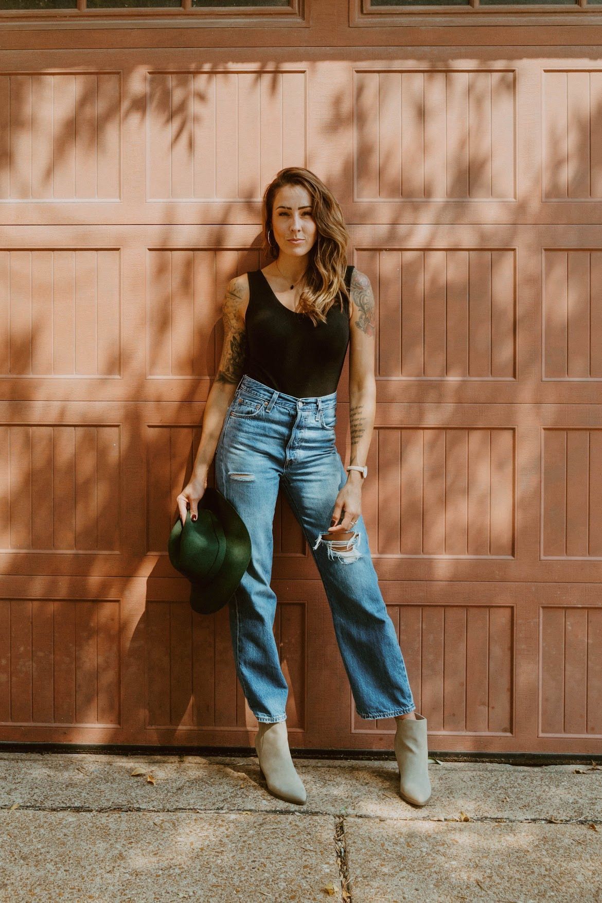Woman leaning against a pink garage door, wearing ripped jeans, black top, and holding a green hat.