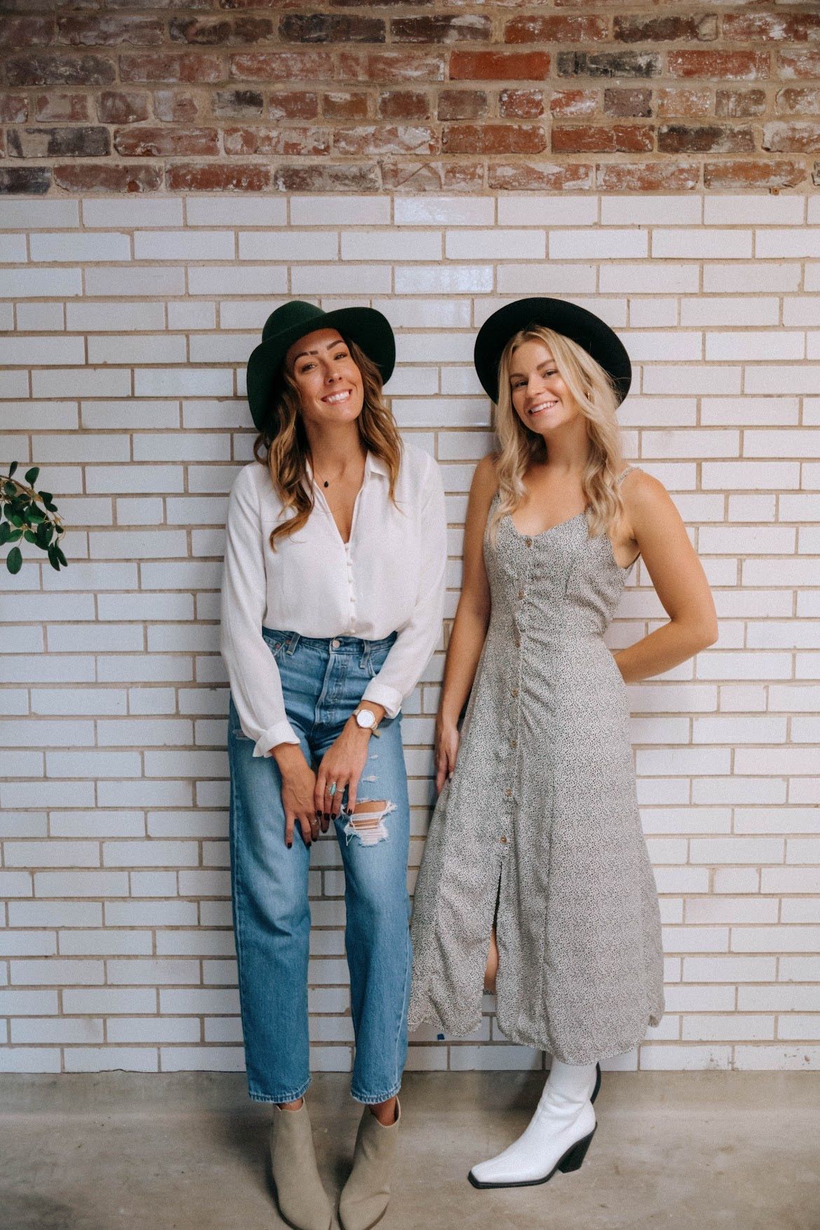 Two women wearing hats pose against a brick wall; one in jeans and a white shirt, the other in a floral dress.