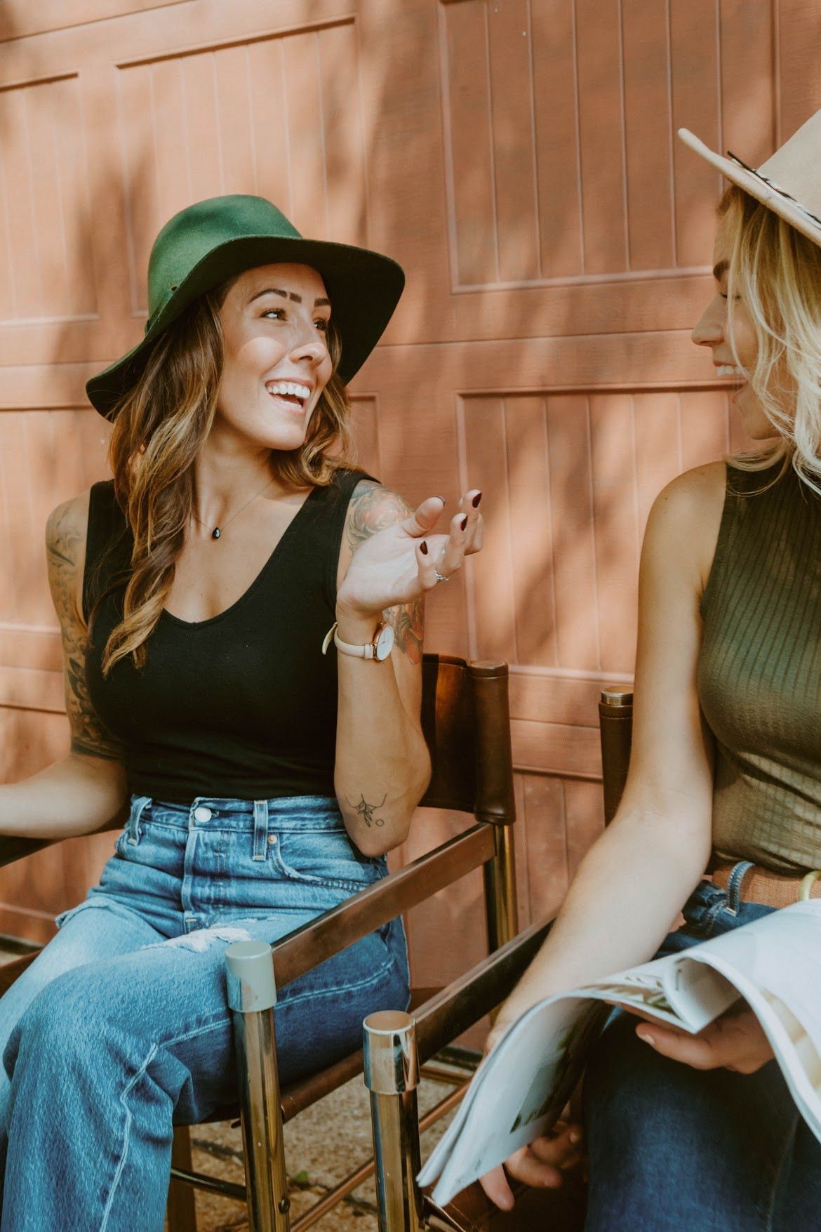 Two women, one in a green hat, laughing while seated in chairs. Jeans, tank tops. Outdoor setting.