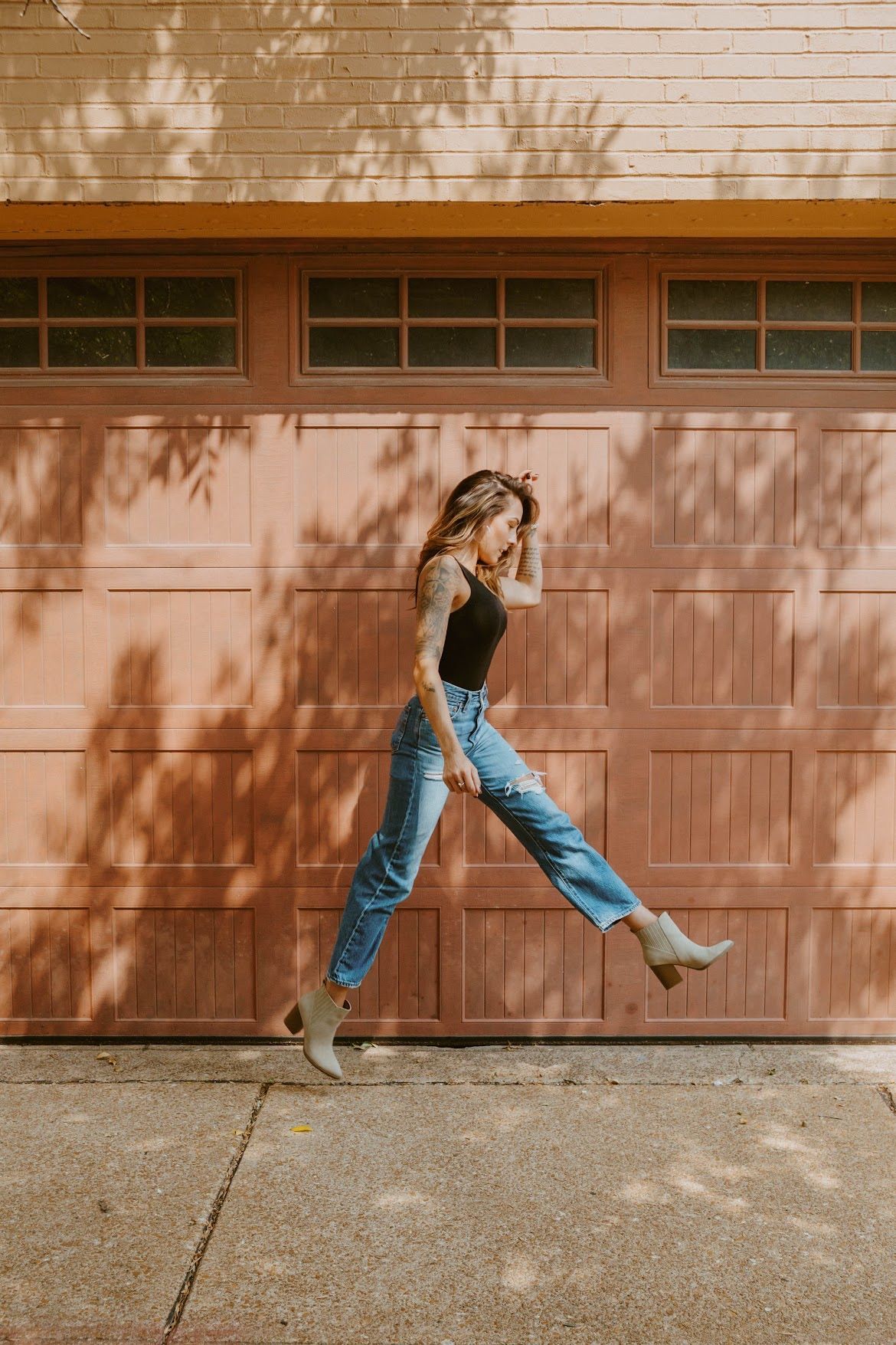 Woman jumping in front of a salmon-colored garage, wearing a black top, jeans, and boots.