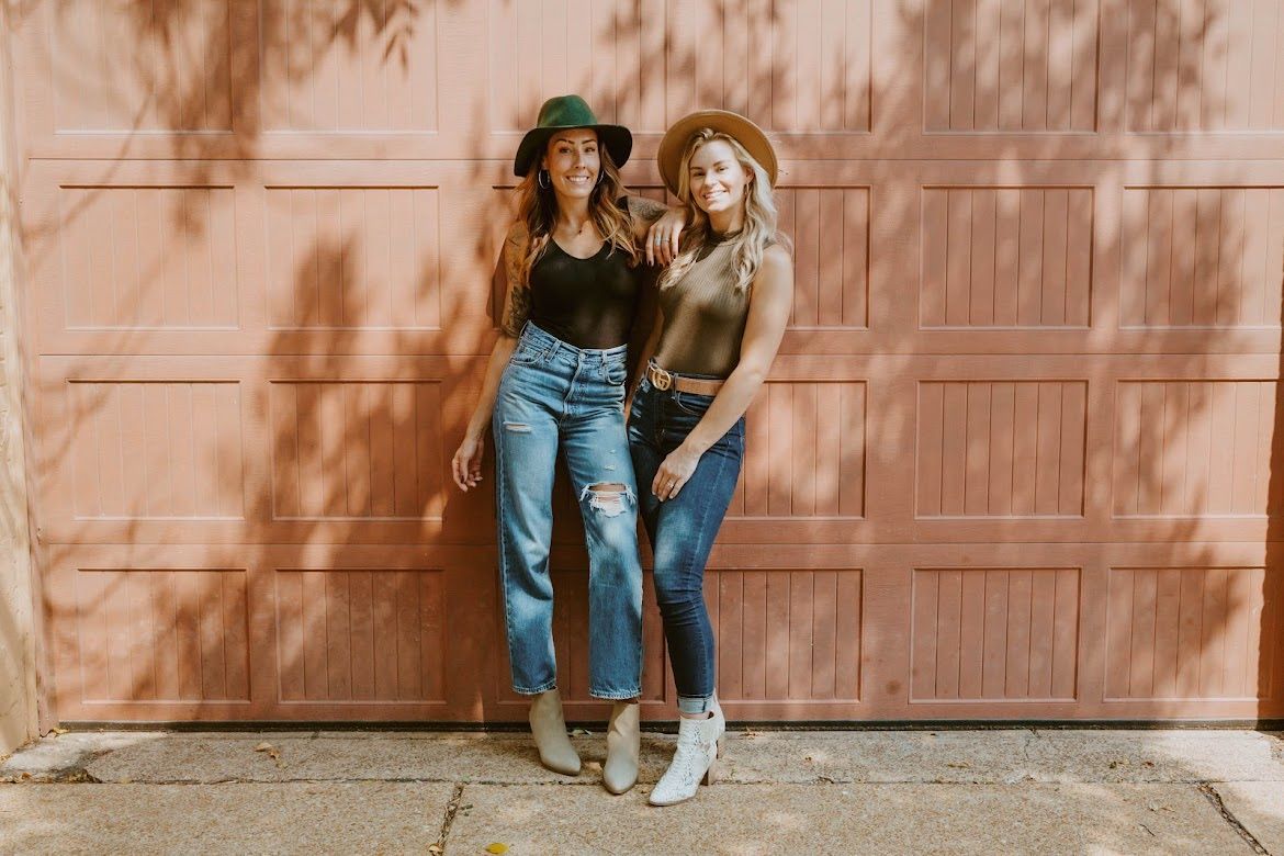 Two women in hats stand by a peach garage door. One wears a black top and jeans, the other an olive top and jeans.