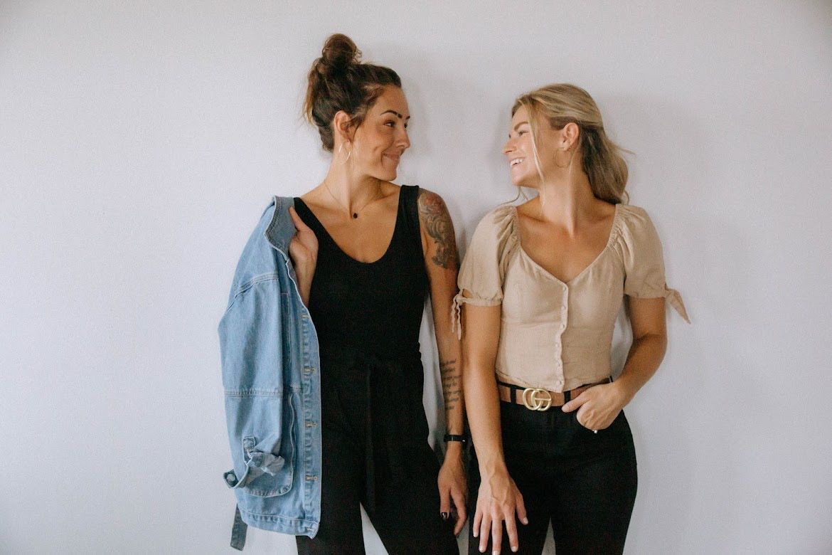 Two women smiling, leaning against a white wall. One in a black jumpsuit with denim jacket, the other in a tan top.