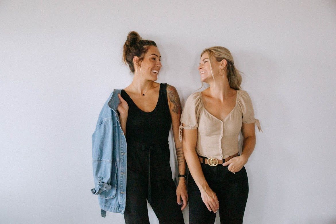 Two women smiling, facing each other against a white wall. One in a black jumpsuit with denim jacket, the other in a tan top and black pants.