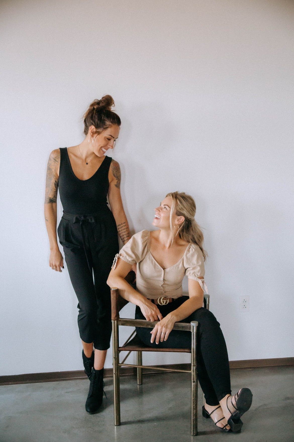 Two women, one standing with arm tattoos, looking at the other seated on a chair. White wall background.