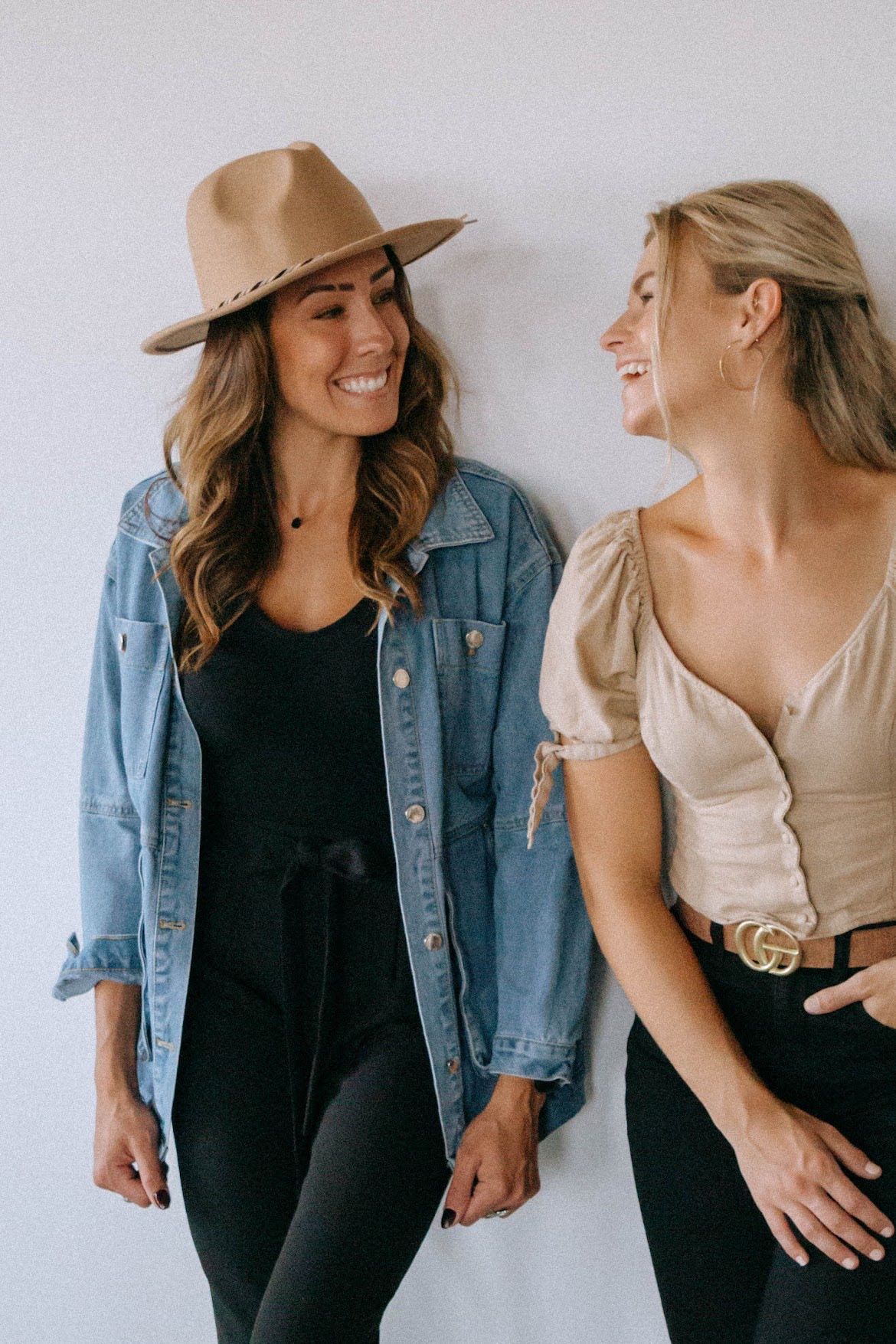 Two smiling women lean against a white wall. One wears a denim jacket and hat, the other a blouse and black pants.