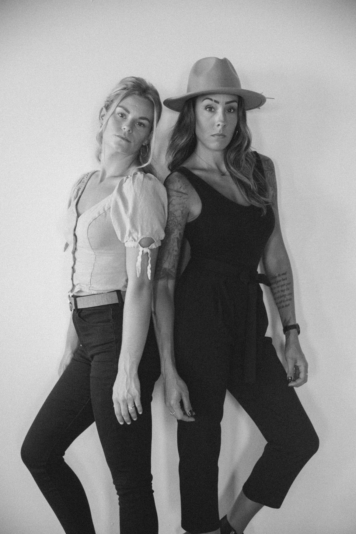 Two people standing back to back, one in a hat and jumpsuit, the other in a blouse and pants, in front of a white wall.