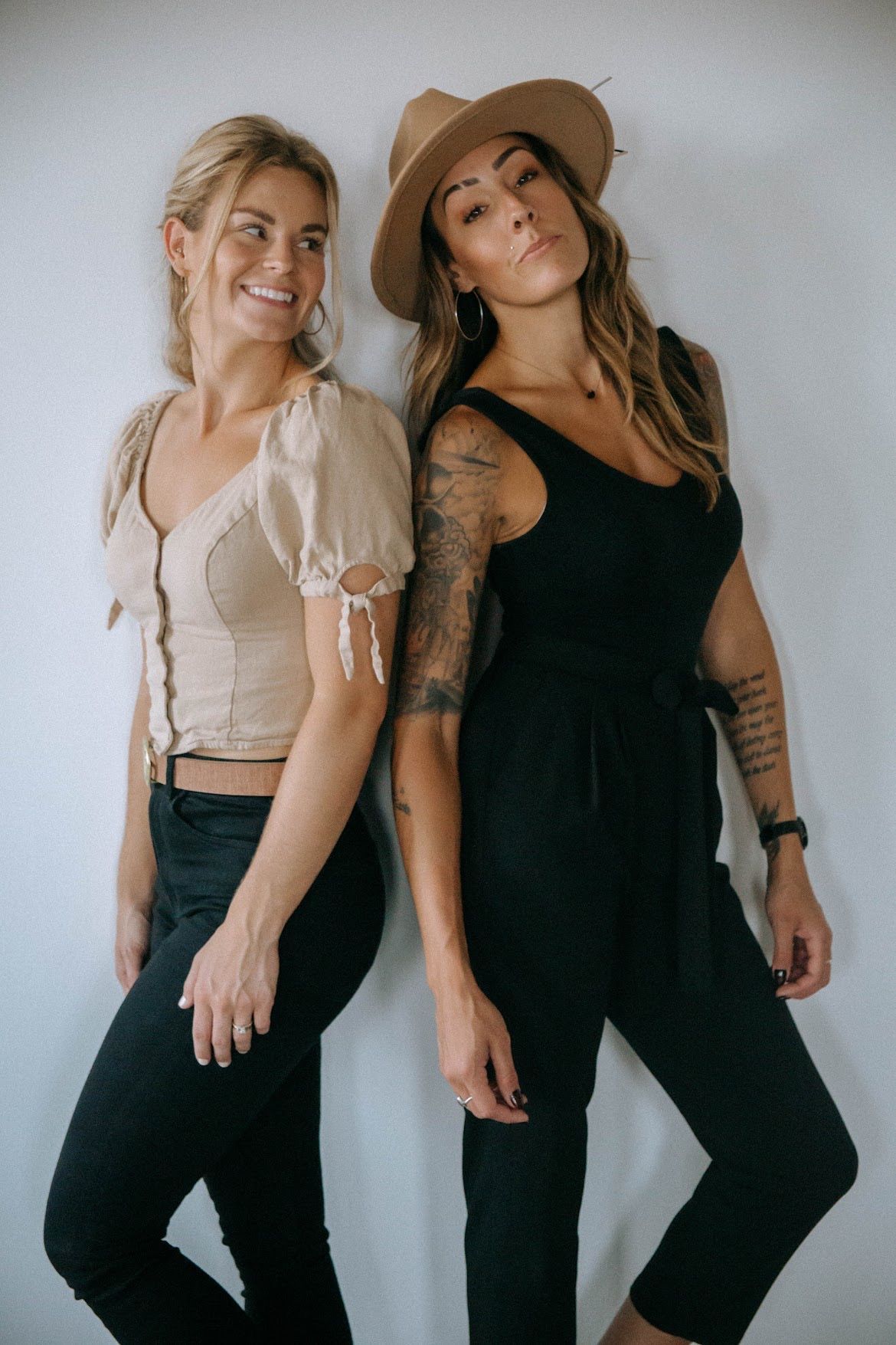 Two women pose against a white wall. One in a tan top and black jeans smiles; the other in a black jumpsuit and hat looks at the camera.