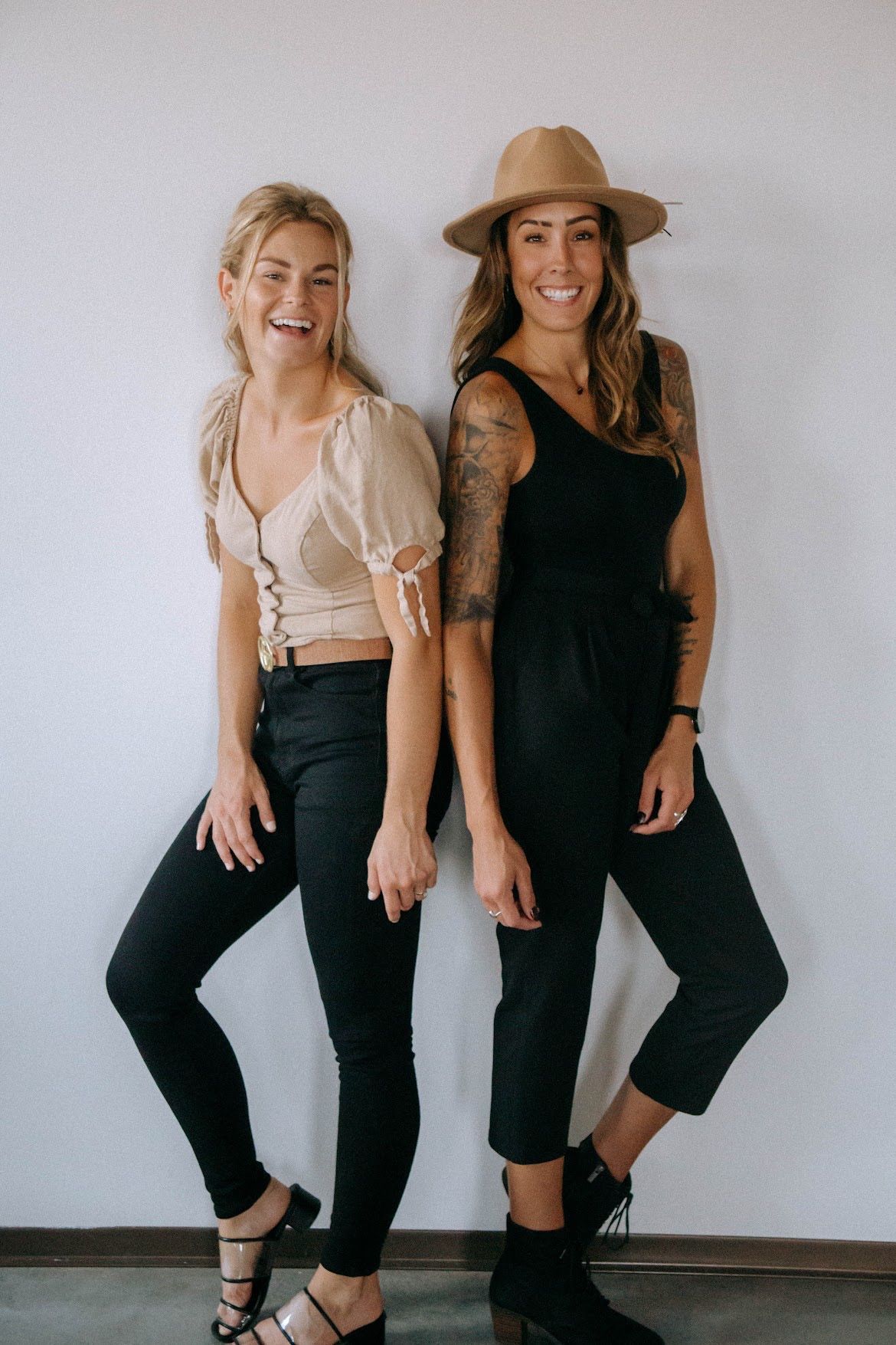 Two women lean against a white wall. One wears a tan top, black pants. The other, black jumpsuit, hat.