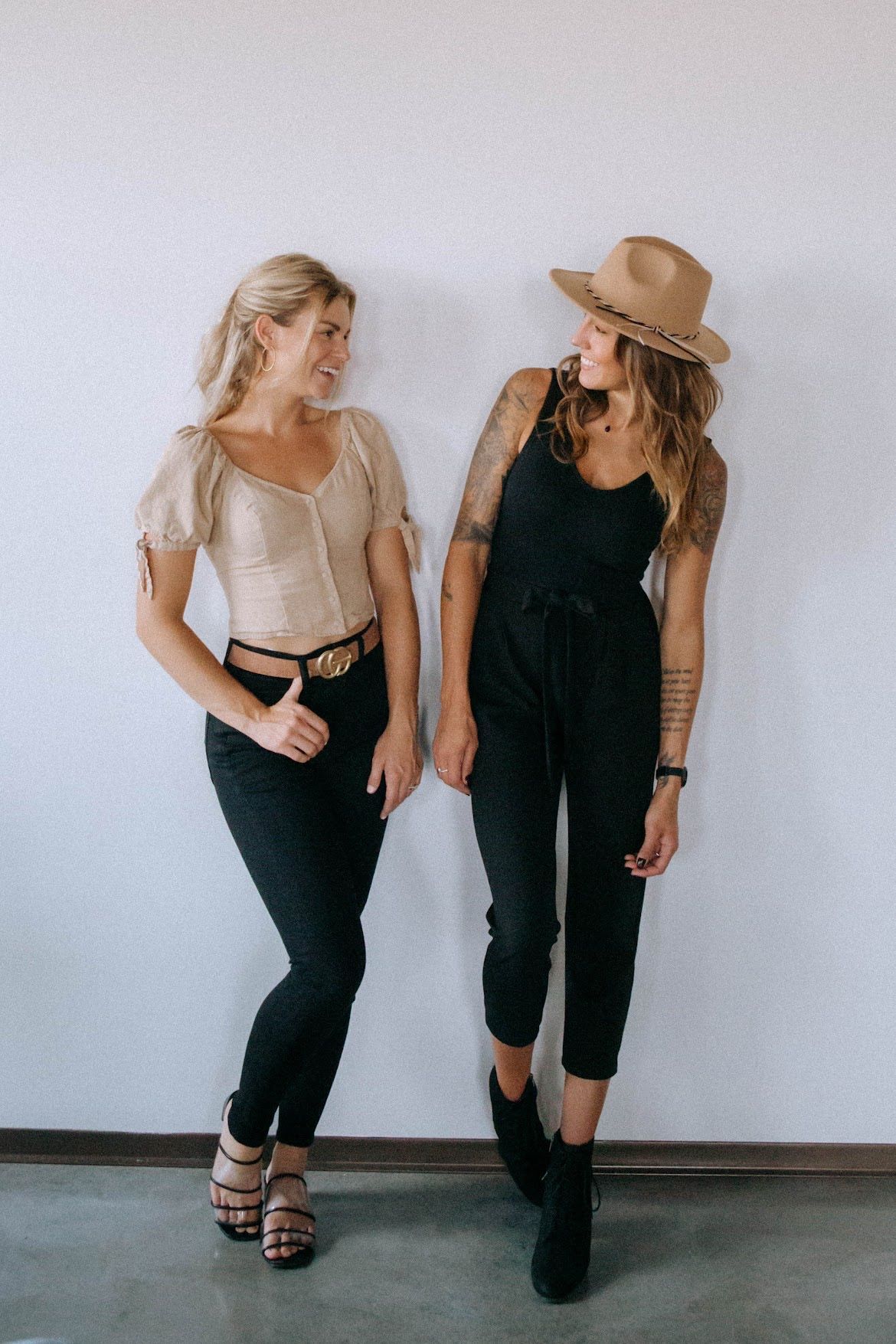 Two women, one in tan top and black pants, the other in black jumpsuit and hat, smile while leaning against a white wall.