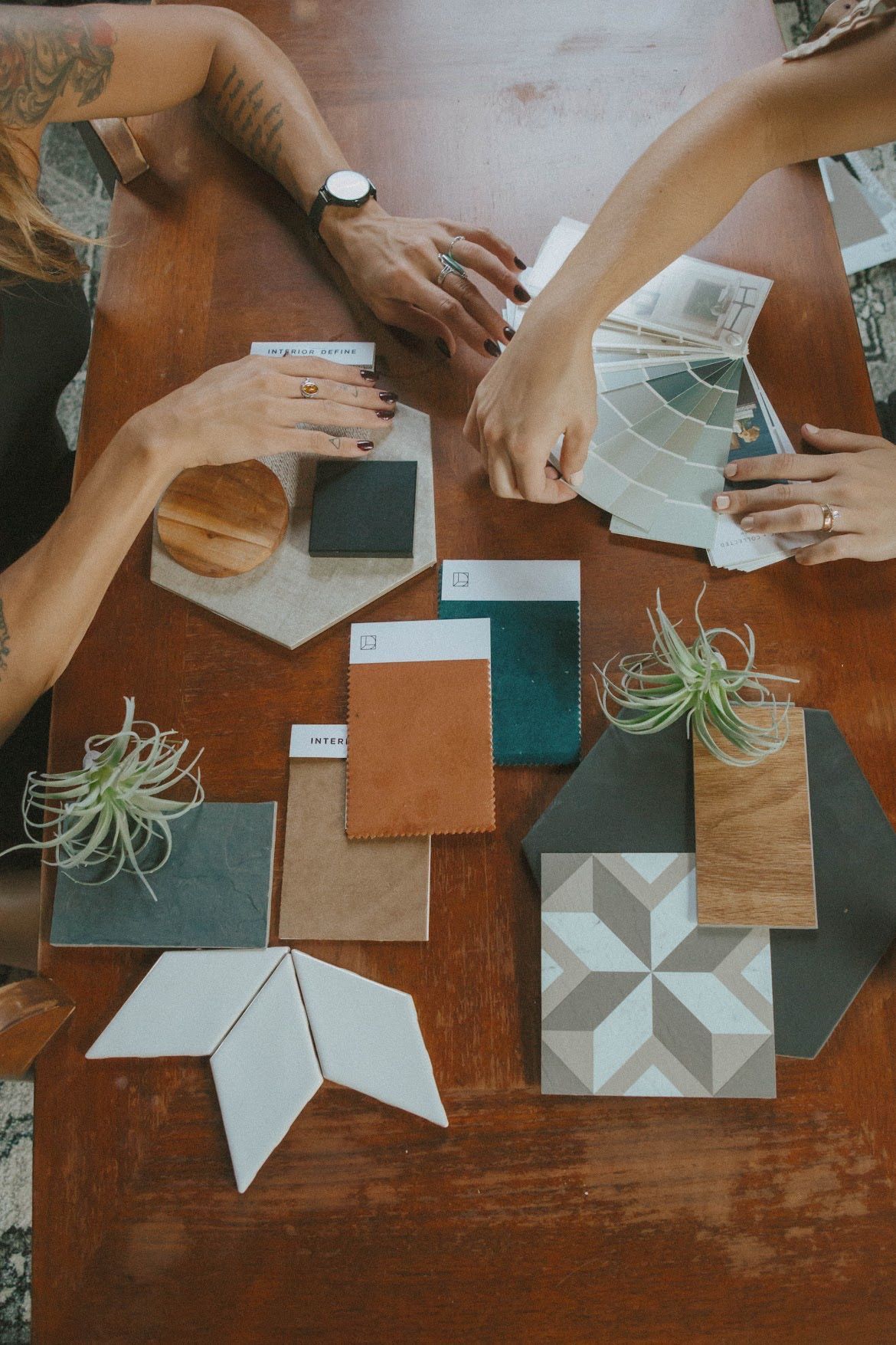 Two people choosing design elements on a table, including tile samples, paint swatches, and small plants.