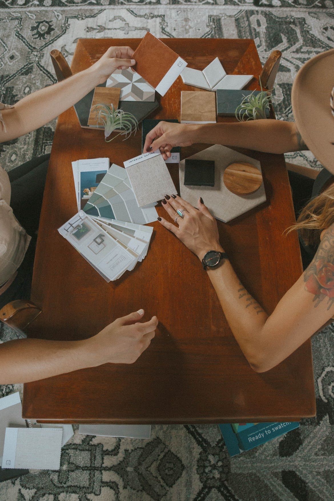 People at a table selecting interior design samples: tiles, paint swatches, wood, and plants.