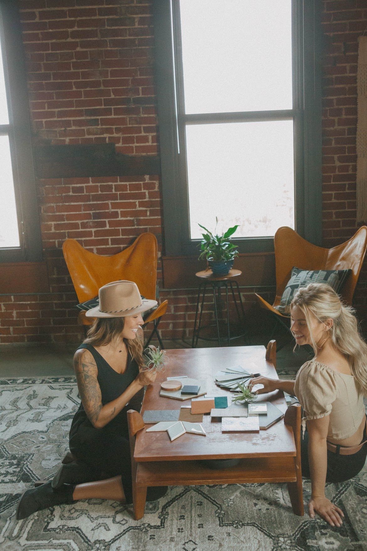 Two people seated at a table with cards; indoor setting with brick wall, butterfly chairs, and large windows.