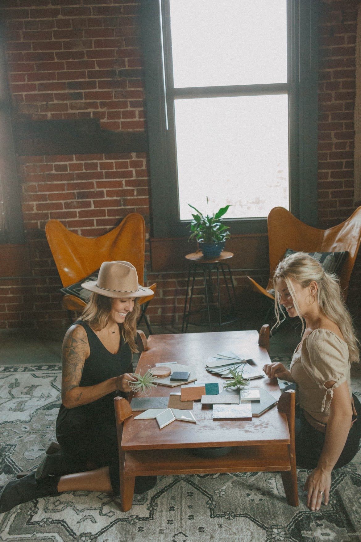 Two people seated around a table, playing cards in a room with a brick wall and natural light.