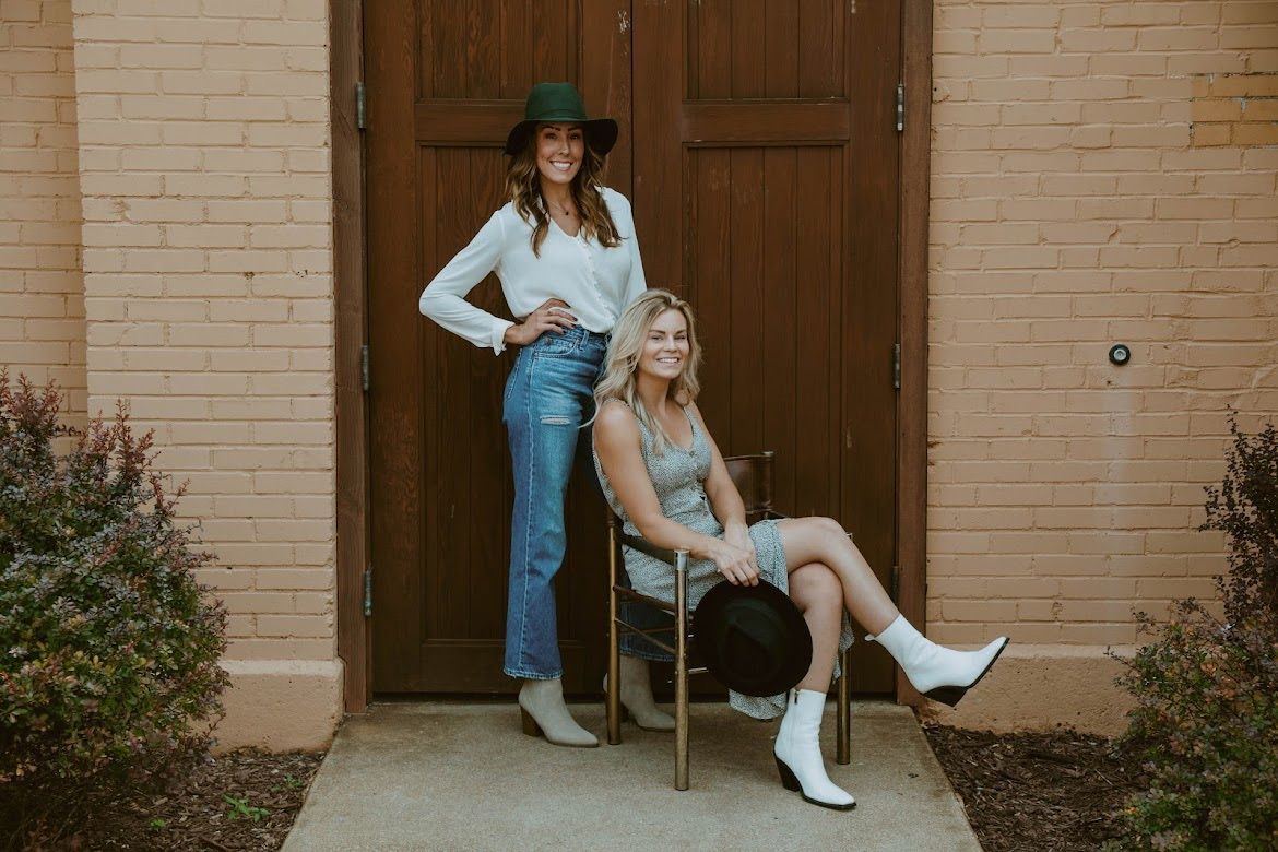 Two women pose outside a brown door. One stands in jeans, the other sits in a dress, both wear hats and boots.