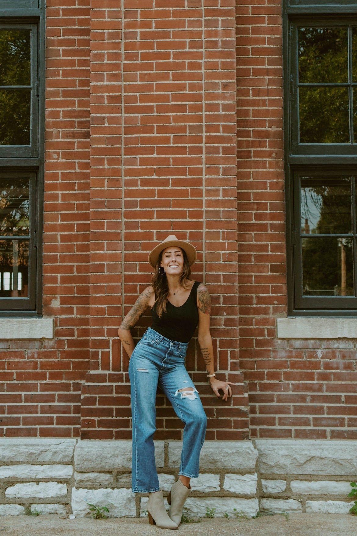 Woman in hat, black top, and jeans leans against a brick wall, smiling.