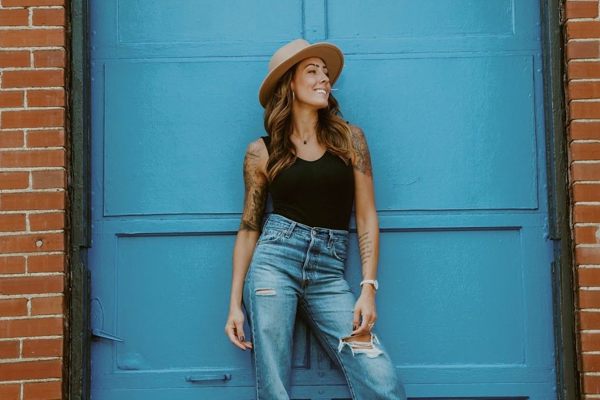 Woman in fedora and jeans, smiling against a blue garage door, arms tattooed.