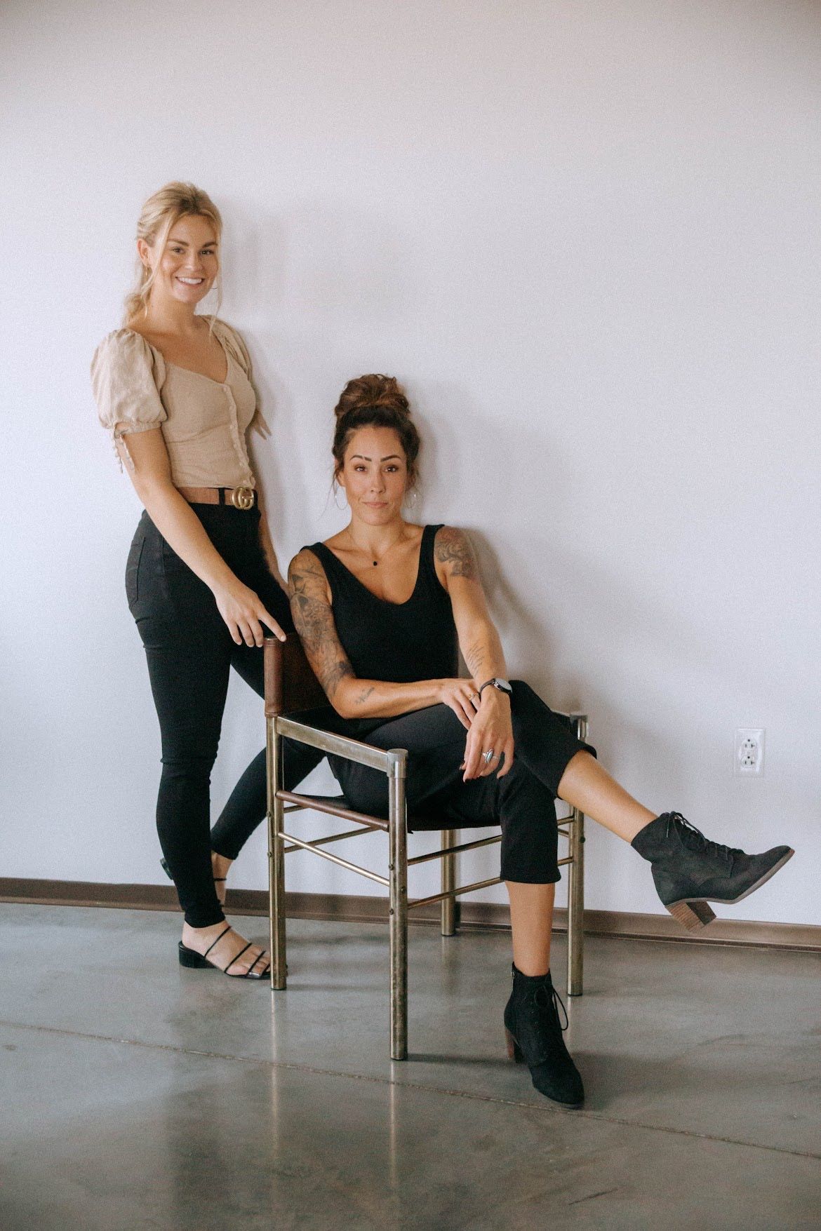 Two women posing: one seated, one standing, leaning against white wall.